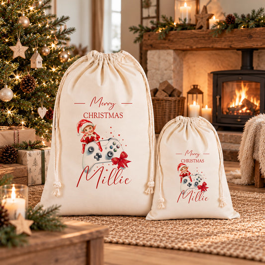 Two white drawstring bags with Christmas-themed designs and text, placed in front of a decorated Christmas tree and a fireplace.