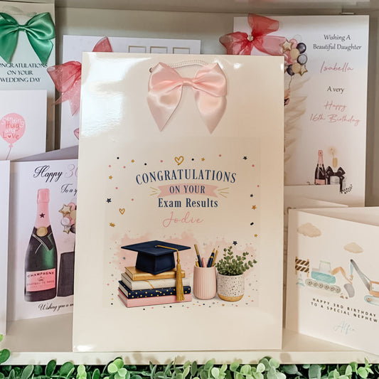 A white congratulations card for exam results is displayed on a shelf among other greeting cards. The main card features a pink bow illustration at the top, the text 'CONGRATULATIONS ON YOUR Exam Results', and an illustration of stacked books, a graduation cap, a pencil cup, and a small plant.