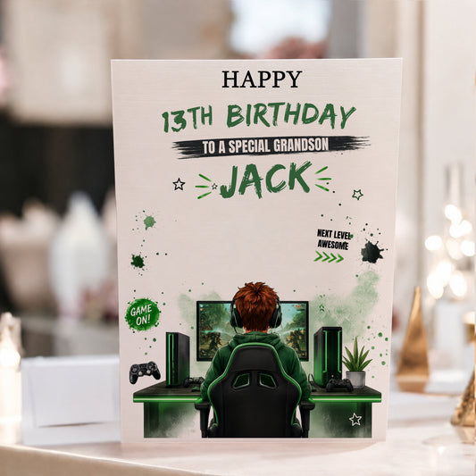 A personalized gaming-themed 13th birthday card for a special grandson, featuring an illustration of a gamer sitting at a desk with a computer setup. The card is displayed upright on a table with a blurred indoor background.