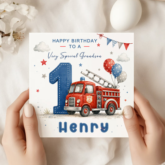 A personalized first birthday card for a grandson, held by two hands. The card features a red fire truck, a large blue number 1, balloons, and the text 'HAPPY BIRTHDAY TO A Very Special Grandson' and 'Henry'.