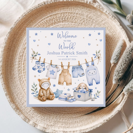 A personalized baby shower sign with a watercolor illustration of baby clothes hanging on a line, a teddy bear, and a bunny in a basket, placed on a woven placemat with dried grasses.