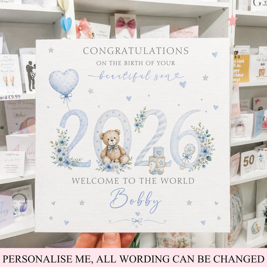 A personalized baby shower card held by a hand in front of a store shelf. The white card features blue and gray text reading 'Congratulations on the birth of your beautiful son 2026 Welcome to the World Bobby' decorated with watercolor-style blue flowers, a teddy bear, and a heart-shaped balloon.