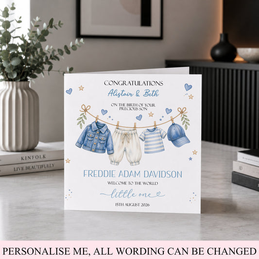 A personalized baby shower congratulations card displayed on a tabletop in a styled home interior. The card features watercolor illustrations of baby clothing hanging on a clothesline, with customizable text for a boy and girl.