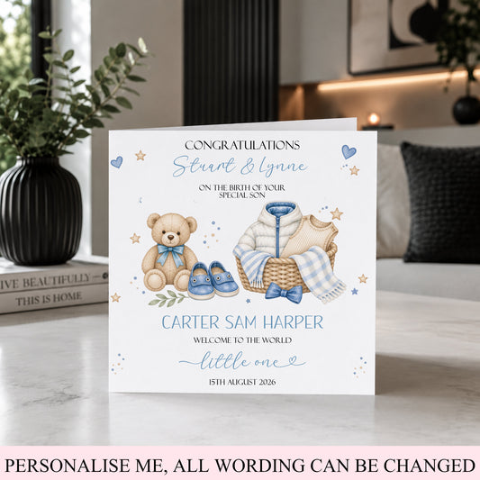 A personalized new baby congratulations card displayed on a marble countertop in a modern kitchen setting. The white card features watercolor illustrations of a teddy bear, a basket with a baby outfit, and blue shoes, with customizable text.