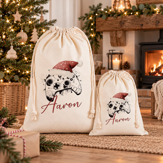 Two white drawstring bags with Christmas-themed designs and the name "Aaron" printed on them, placed on a wooden floor in front of a decorated Christmas tree.