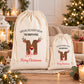 Two white drawstring bags with Christmas-themed designs and text, placed on a wooden surface with pine cones and other holiday decorations in the background.