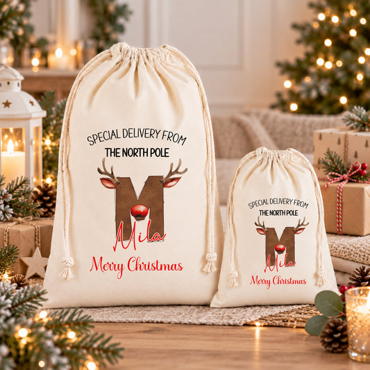 Two white drawstring bags with Christmas-themed designs and text, placed on a wooden surface with pine cones and other holiday decorations in the background.
