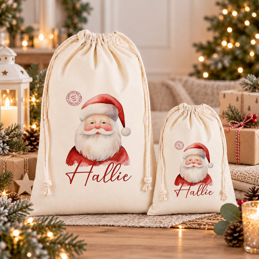 two white drawstring bags with a Santa Claus design, placed on a wooden surface. The bags are personalized with the name "Hallie" printed on them. The background of the image is decorated with Christmas lights and pine cones, creating a festive atmosphere.