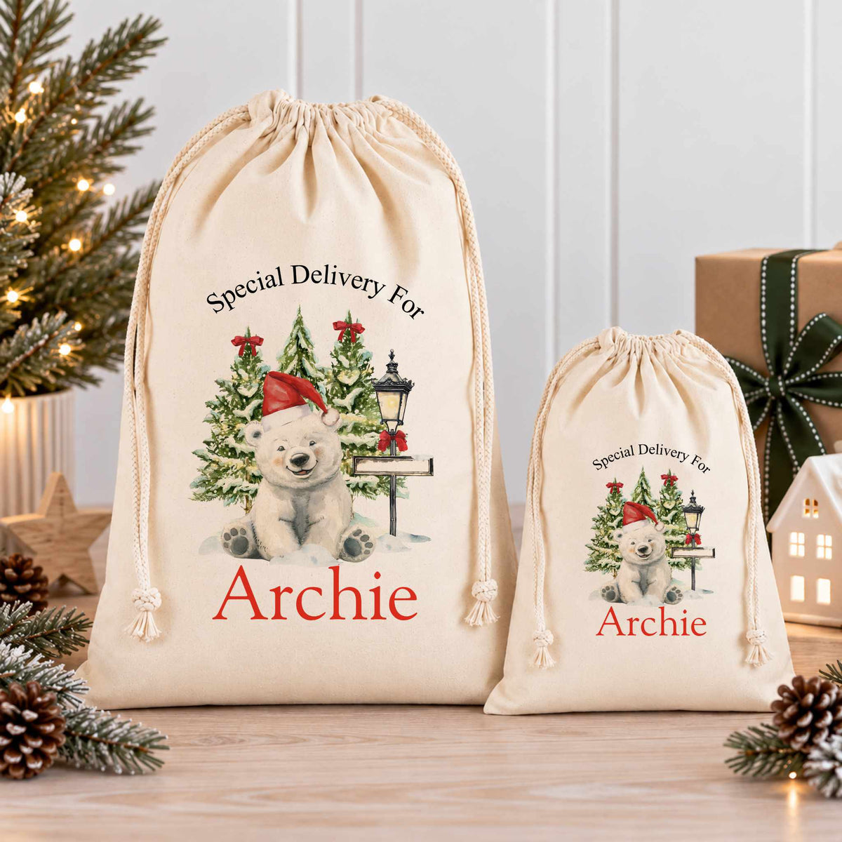 Two white drawstring bags with Christmas-themed illustrations and the name "Archie" printed on them, placed on a wooden surface with pine cones and a small Christmas tree in the background.