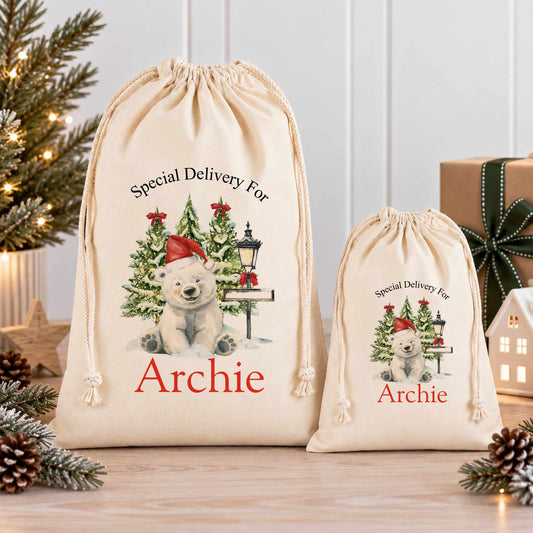 Two white drawstring bags with Christmas-themed illustrations and the name "Archie" printed on them, placed on a wooden surface with pine cones and a small Christmas tree in the background.