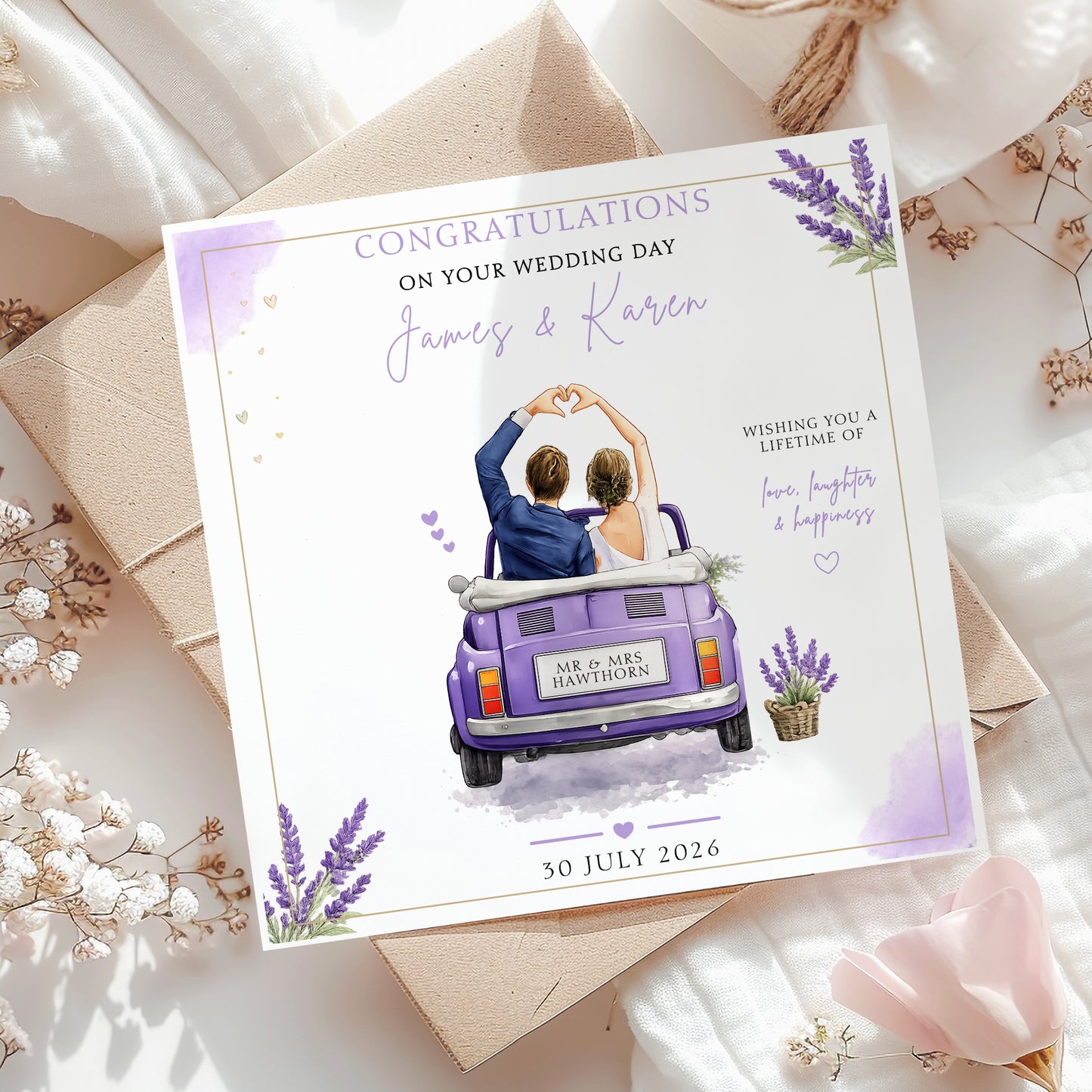 A personalized wedding congratulations card featuring an illustration of a couple in a purple van, styled on top of kraft envelopes and white fabric with dried flowers.