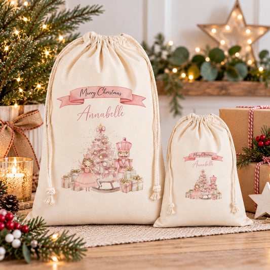 two white drawstring bags with Christmas-themed illustrations and personalized names on them, placed on a wooden surface with a Christmas tree and other holiday decorations in the background.