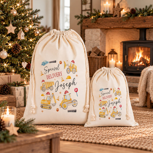 Two white drawstring bags with illustrations of construction vehicles and the text "Special Delivery Joseph" printed on them, placed in front of a Christmas tree and a fireplace.