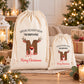 Two white drawstring bags with Christmas-themed designs and text, placed on a wooden surface with Christmas decorations and lights in the background.