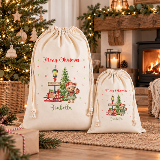 Two white drawstring bags with Christmas-themed illustrations and personalized names are displayed in front of a decorated Christmas tree.