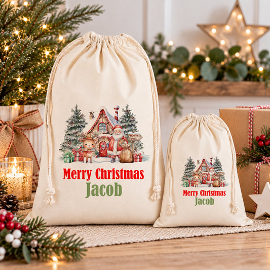 two Christmas-themed drawstring bags, one larger and one smaller, both with a festive illustration of Santa Claus and Christmas trees. The larger bag is labeled "Merry Christmas Jacob" and the smaller bag is labeled "Merry Christmas Jacob". The bags are placed on a wooden surface, surrounded by holiday decorations such as a Christmas tree, a star, and other festive items.
