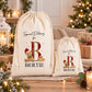 two white drawstring bags with a teddy bear design and the letter "B" printed on them, placed on a table with Christmas decorations and gifts.