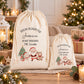 Two white drawstring bags with Christmas-themed illustrations and text, placed on a table with holiday decorations and gifts.