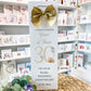A personalized wedding anniversary gift bag standing upright on a woven straw mat, featuring a large gold bow at the top and elegant gold and black text celebrating a 30th wedding anniversary. The background shows shelves filled with greeting cards.