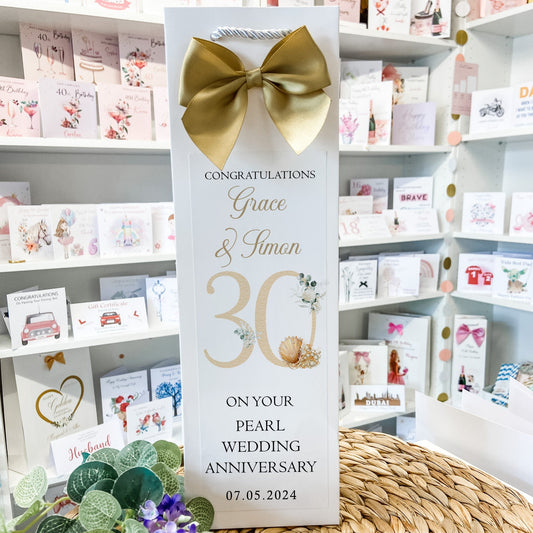 A personalized wedding anniversary gift bag standing upright on a woven straw mat, featuring a large gold bow at the top and elegant gold and black text celebrating a 30th wedding anniversary. The background shows shelves filled with greeting cards.