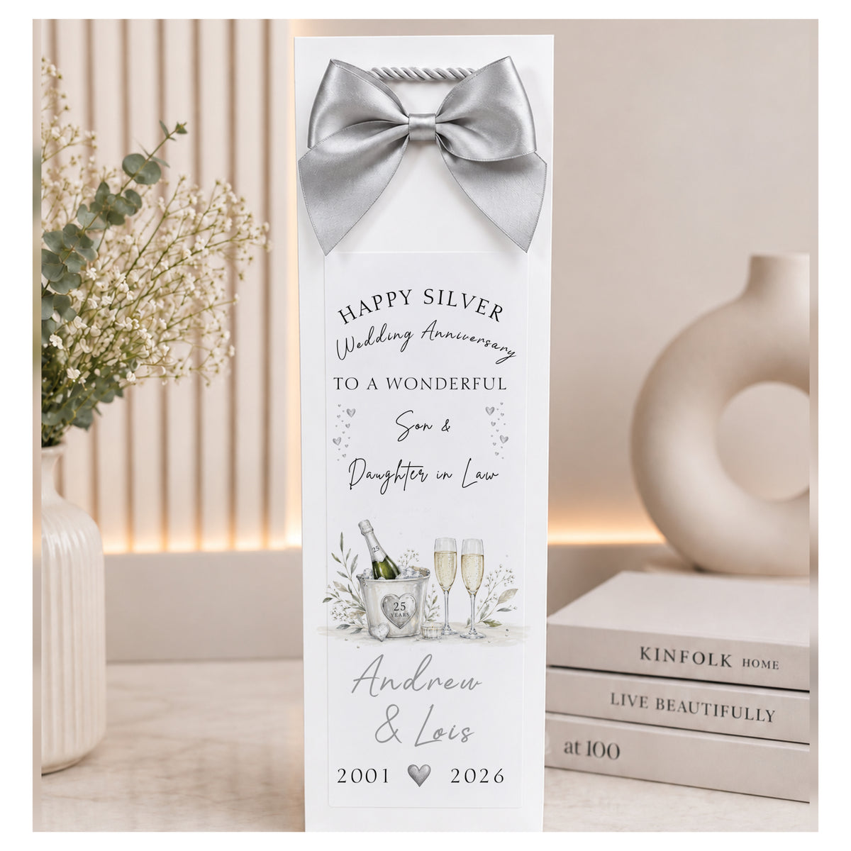 A personalized silver-toned champagne bottle gift tag with a matching satin bow, featuring a custom 'Happy Silver Wedding Anniversary' message for Andrew & Lois. The tag is styled on a tabletop next to a vase of baby's breath and a stack of books.