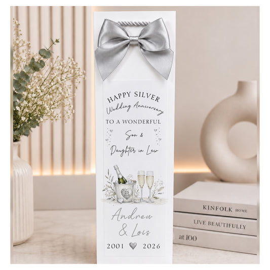 A personalized silver-toned champagne bottle gift tag with a matching satin bow, featuring a custom 'Happy Silver Wedding Anniversary' message for Andrew & Lois. The tag is styled on a tabletop next to a vase of baby's breath and a stack of books.