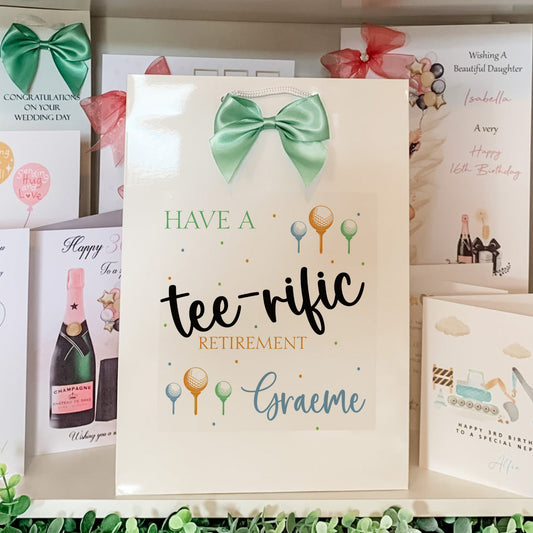 Greeting card with a green bow and text 'Have a tee-rific retirement, Graeme' on a shelf with other cards.