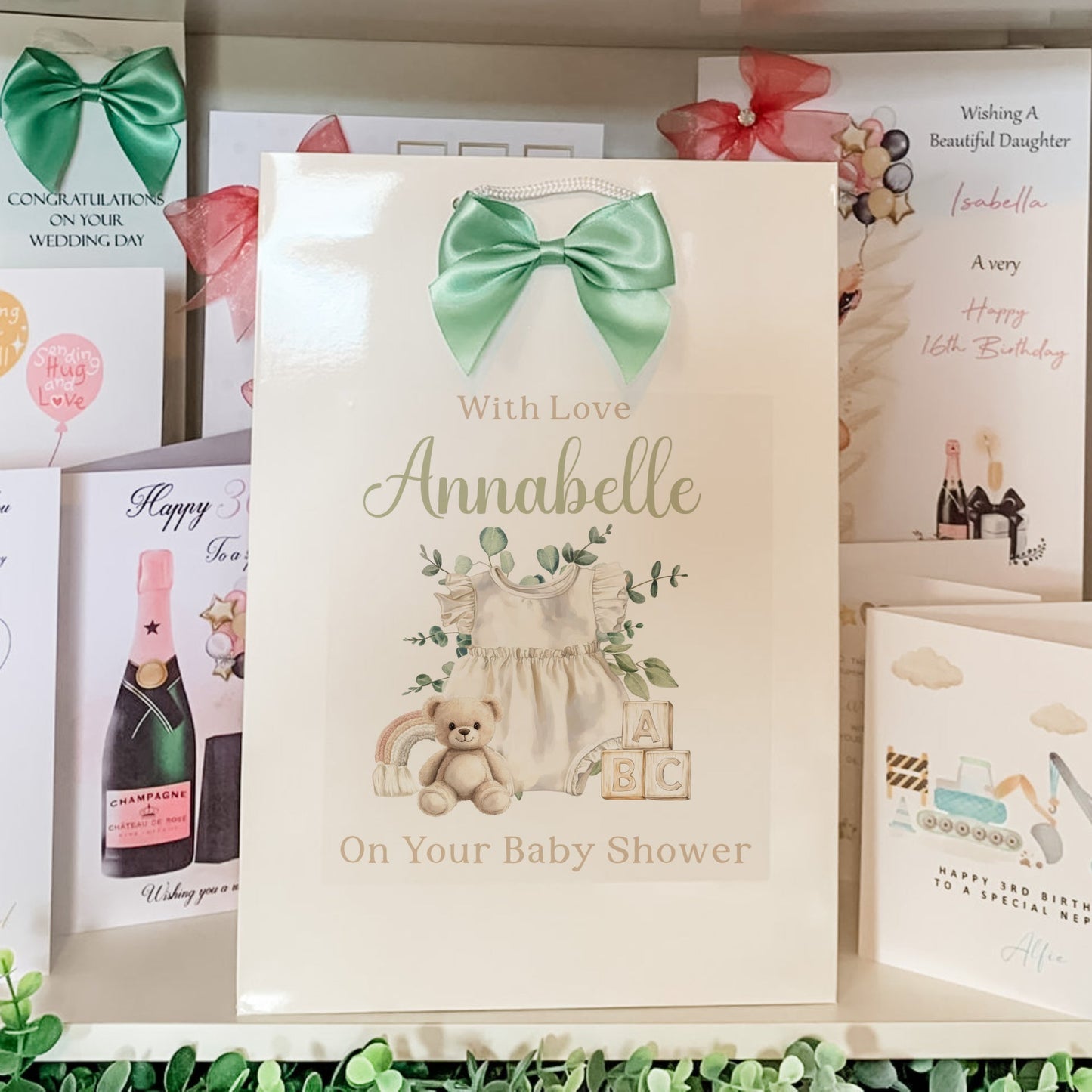 Baby shower card with a green bow, teddy bear, and baby outfit on a shelf with other cards.