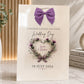 A cream-colored wedding gift bag featuring a purple bow at the top and a watercolor floral wreath with the names 'Louis & Sophia' and the date '18 JULY 2026'. The bag is styled on a marble surface next to a vase with greenery and a stack of books.