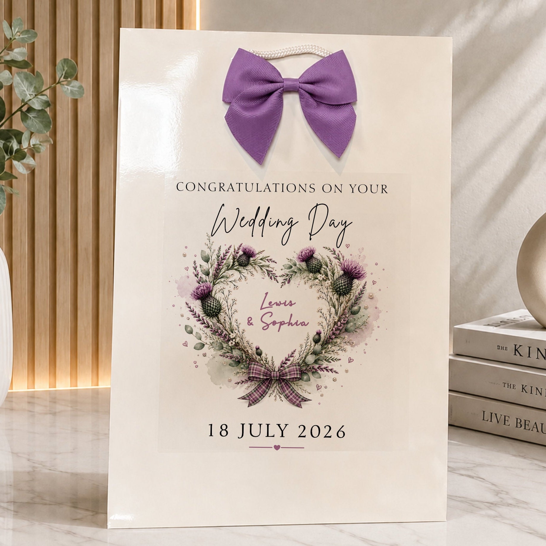 A cream-colored wedding gift bag featuring a purple bow at the top and a watercolor floral wreath with the names 'Louis & Sophia' and the date '18 JULY 2026'. The bag is styled on a marble surface next to a vase with greenery and a stack of books.
