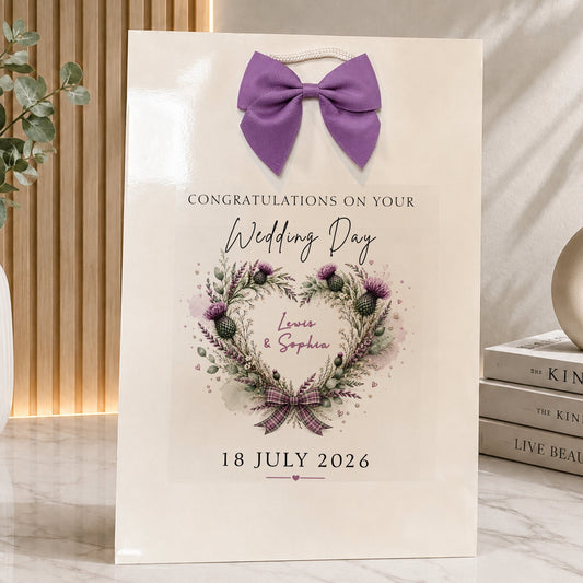 A cream-colored wedding gift bag featuring a purple bow at the top and a watercolor floral wreath with the names 'Louis & Sophia' and the date '18 JULY 2026'. The bag is styled on a marble surface next to a vase with greenery and a stack of books.