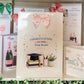 A white congratulations card for exam results is displayed on a shelf among other greeting cards. The main card features a pink bow illustration at the top, the text 'CONGRATULATIONS ON YOUR Exam Results', and an illustration of stacked books, a graduation cap, a pencil cup, and a small plant.