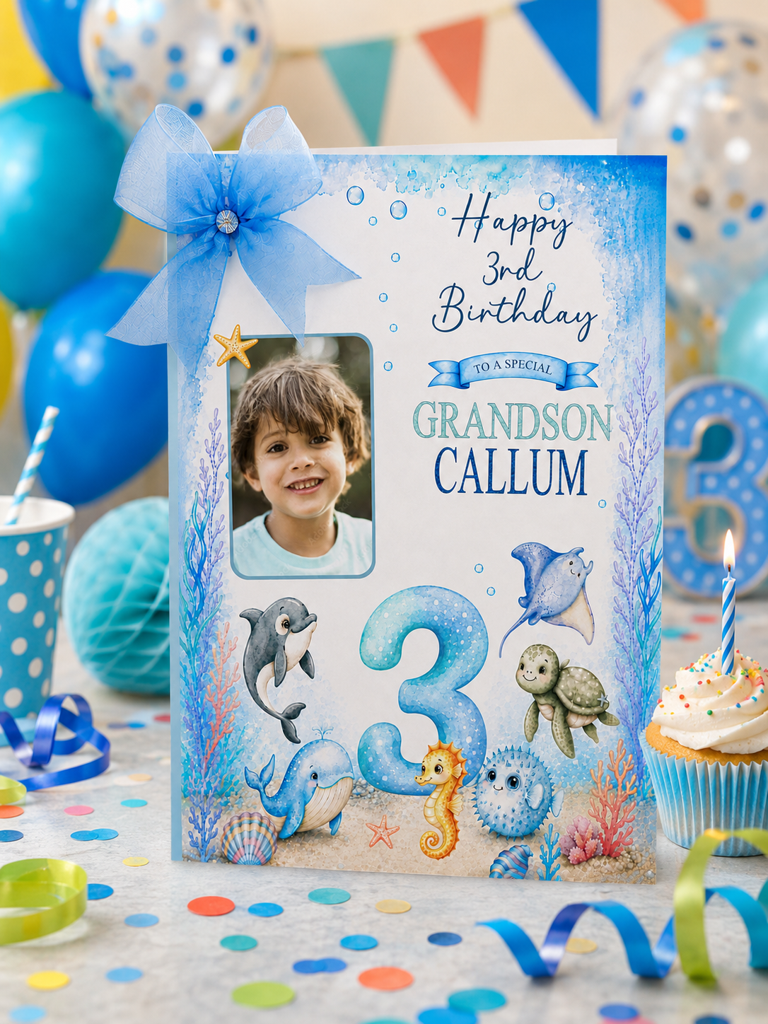 A personalized 3rd birthday card for 'Grandson Callum' with an underwater theme, styled on a party table with balloons, a cup, a cupcake, and confetti. The card features a blue ribbon bow, a photo of the grandson, and illustrations of dolphins, a seahorse, and a turtle.