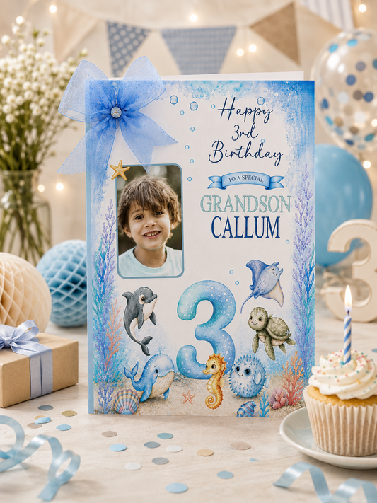 A personalized underwater-themed 3rd birthday photo card for 'Grandson Callum' is displayed on a decorated birthday table alongside a cupcake, gift box, and balloons.