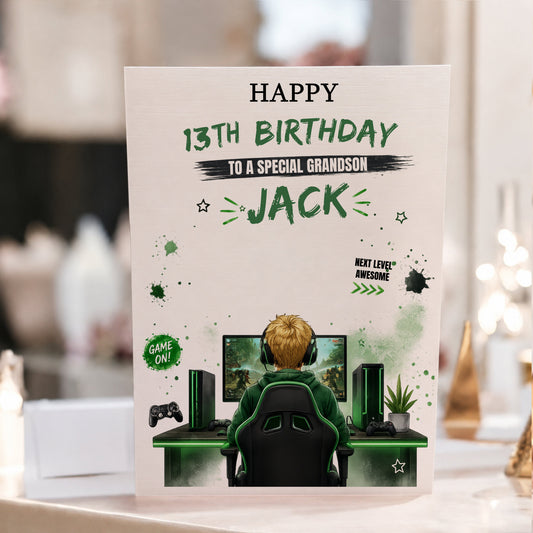 A personalized gaming-themed 13th birthday card for a special grandson, featuring a cartoon illustration of a gamer sitting at a desk with a computer setup. The card is displayed upright on a table with a blurred indoor background.