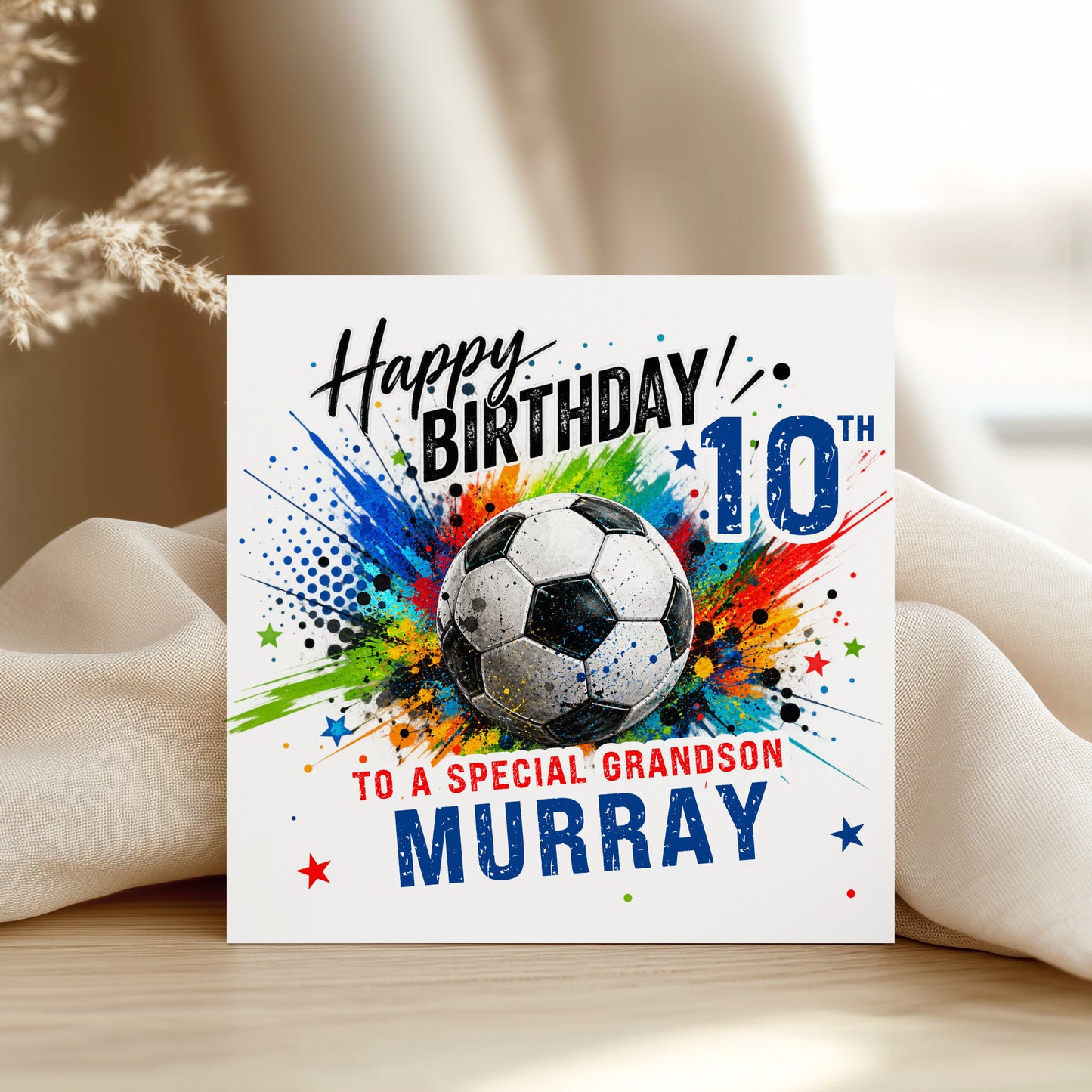 A square birthday card with a soccer ball graphic and colorful paint splashes, reading 'Happy Birthday! any age any name and relationship displayed on a light surface with draped fabric in the background.