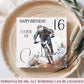 A square personalized birthday card for a mountain biker, displayed on a woven straw mat with dried grass stems. The card features a photo of a cyclist and customizable text reading 'HAPPY BIRTHDAY TO A SPECIAL SON', '16', and 'Calvin'.