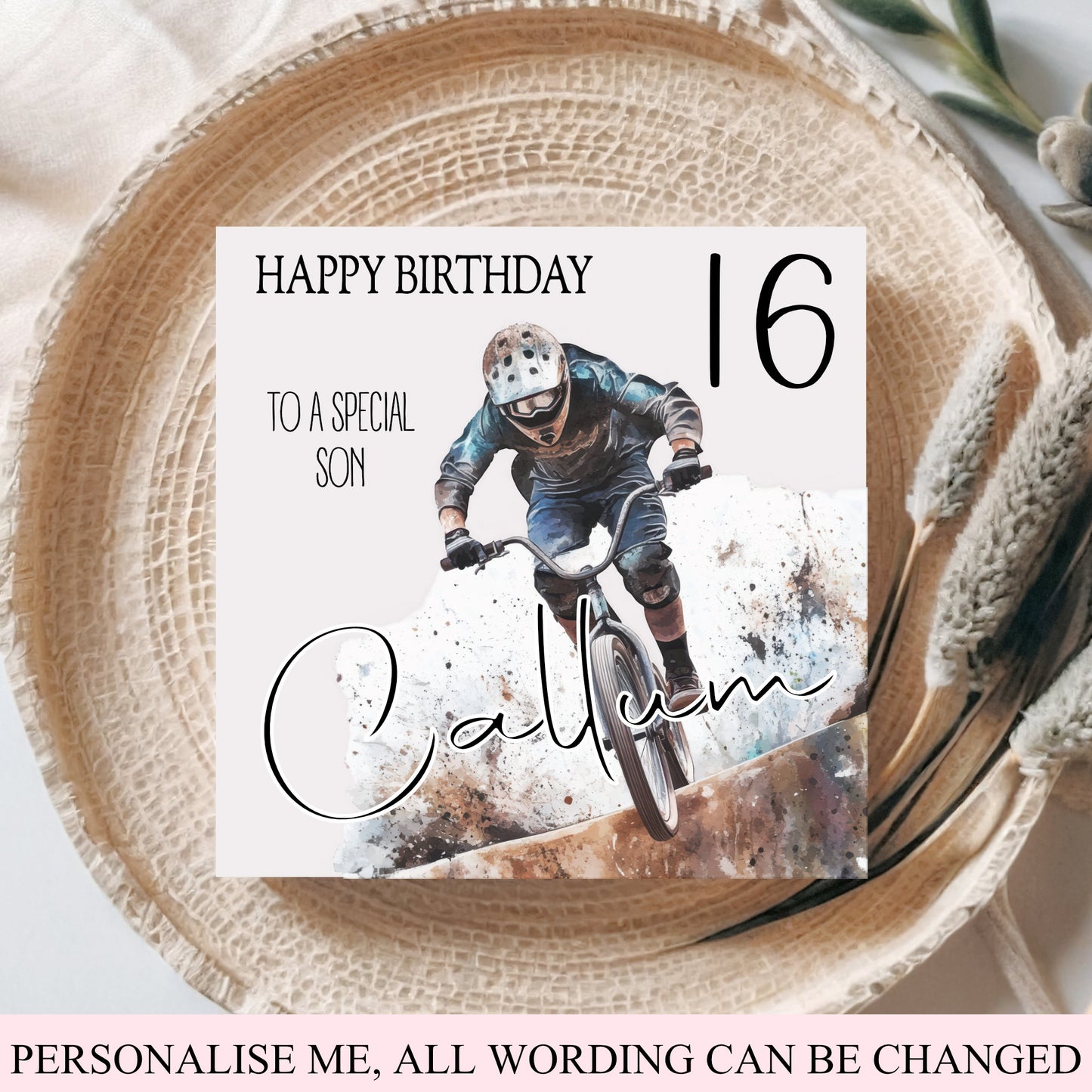 A square personalized birthday card for a mountain biker, displayed on a woven straw mat with dried grass stems. The card features a photo of a cyclist and customizable text reading 'HAPPY BIRTHDAY TO A SPECIAL SON', '16', and 'Calvin'.