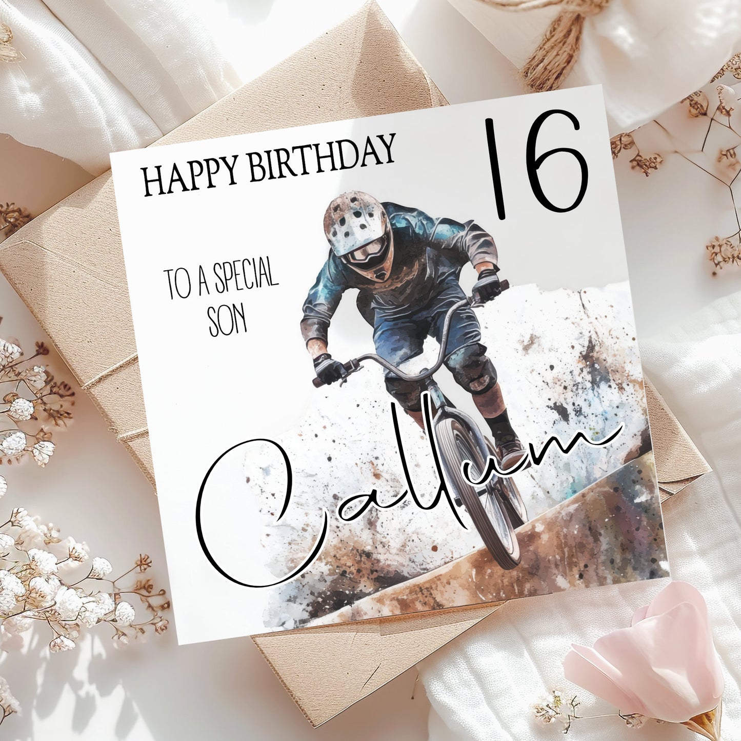 A square birthday card for a 16th birthday son, styled in a flat-lay arrangement on brown kraft paper envelopes and white fabric with dried flowers. The card features a photo of a mountain biker and the text 'HAPPY BIRTHDAY TO A SPECIAL SON', '16', and 'Calvin' in script.