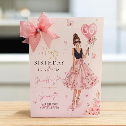 A pink birthday card addressed to a special granddaughter, featuring an illustration of a woman in a floral pink dress holding a heart-shaped balloon with the number 50. The card is decorated with a large pink bow and stands on a light wooden surface.