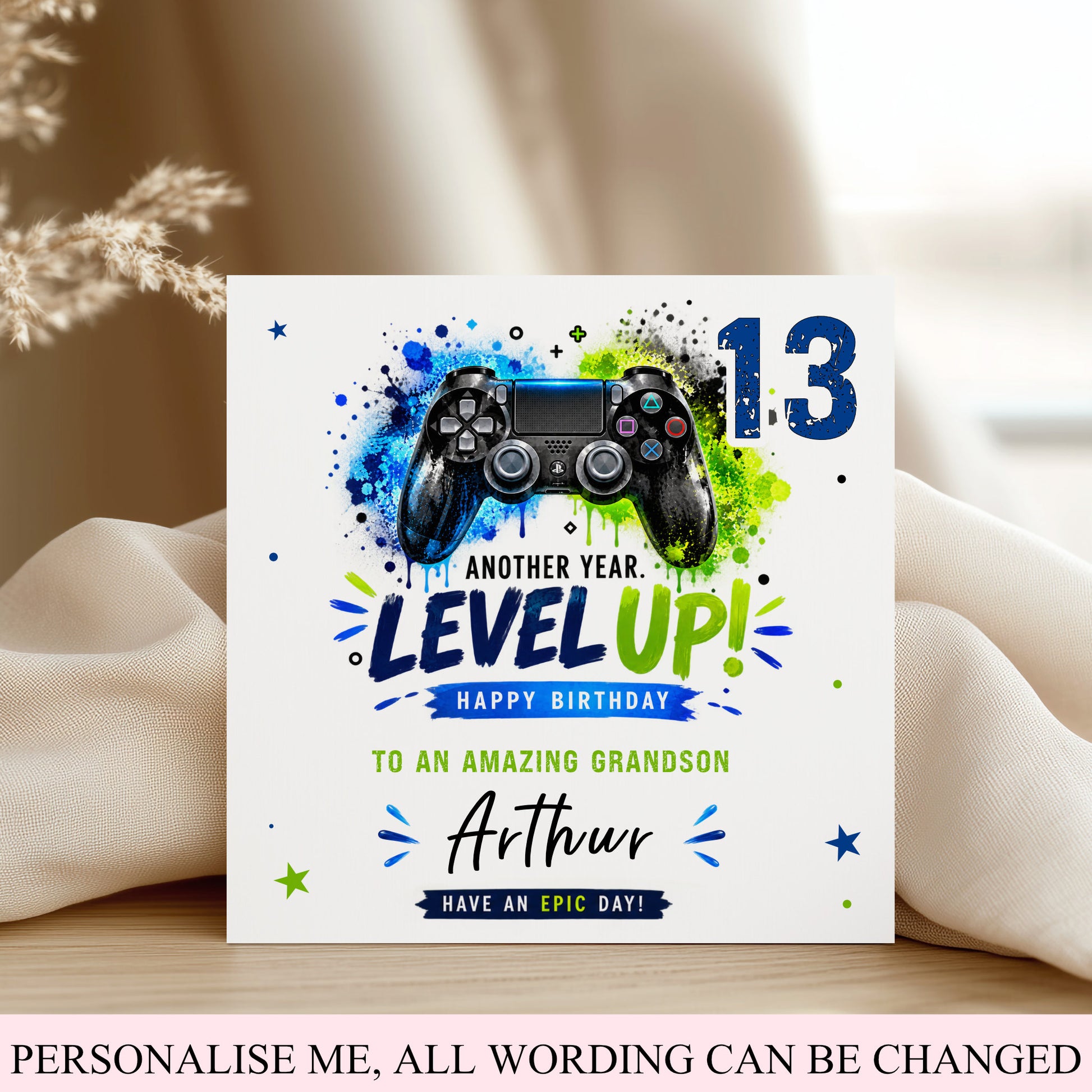 A square birthday card for a 13th birthday grandson, featuring a black video game controller graphic with blue and green paint splatters, the number 13, and the text 'LEVEL UP! HAPPY BIRTHDAY TO AN AMAZING GRANDSON Arthur'. The card is displayed on a wooden surface with beige fabric and dried grass in the background.