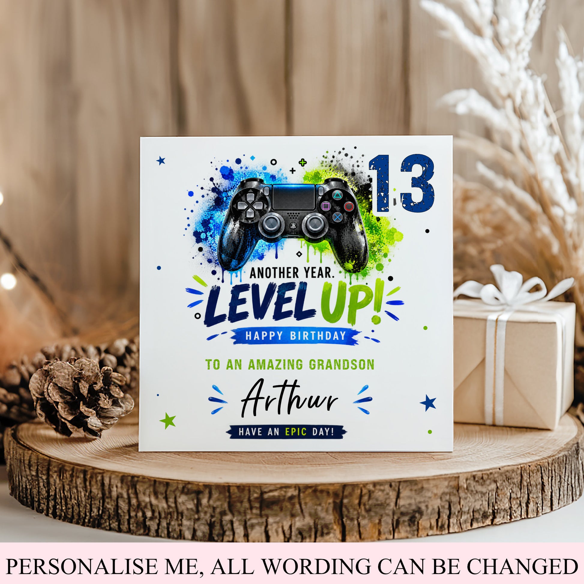 A custom square birthday card styled on a rustic wooden slice with a pinecone and a wrapped gift box. The card features a video game controller graphic over a blue, green, and black paint splatter design, with the text 'ANOTHER YEAR.