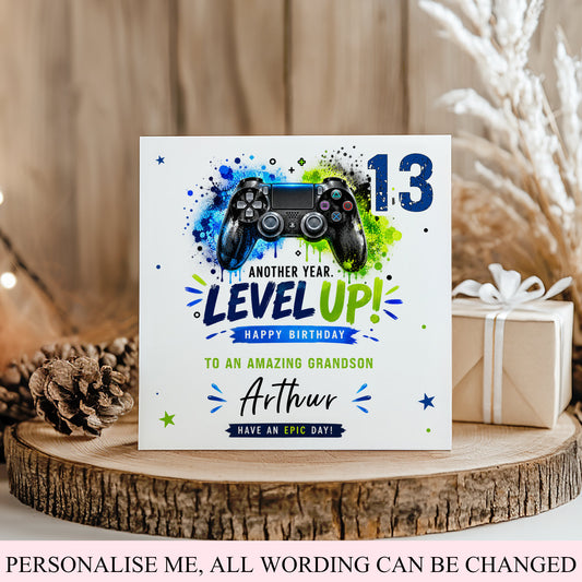 A custom square birthday card styled on a rustic wooden slice with a pinecone and a wrapped gift box. The card features a video game controller graphic over a blue, green, and black paint splatter design, with the text 'ANOTHER YEAR.