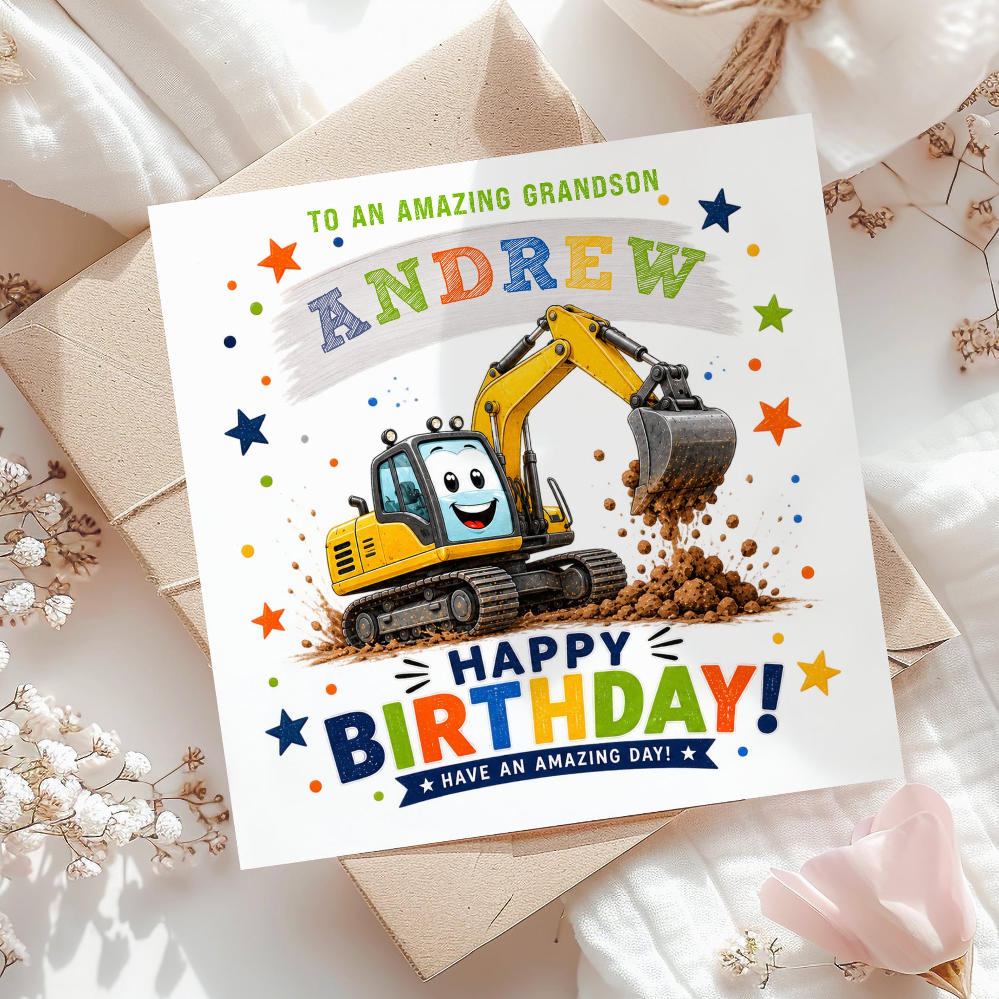 A square birthday card for a grandson, featuring a cartoon excavator digging dirt and the text 'TO AN AMAZING GRANDSON ANDREW HAPPY BIRTHDAY! HAVE AN AMAZING DAY!'. The card is styled on top of kraft paper envelopes and white fabric with dried flowers.