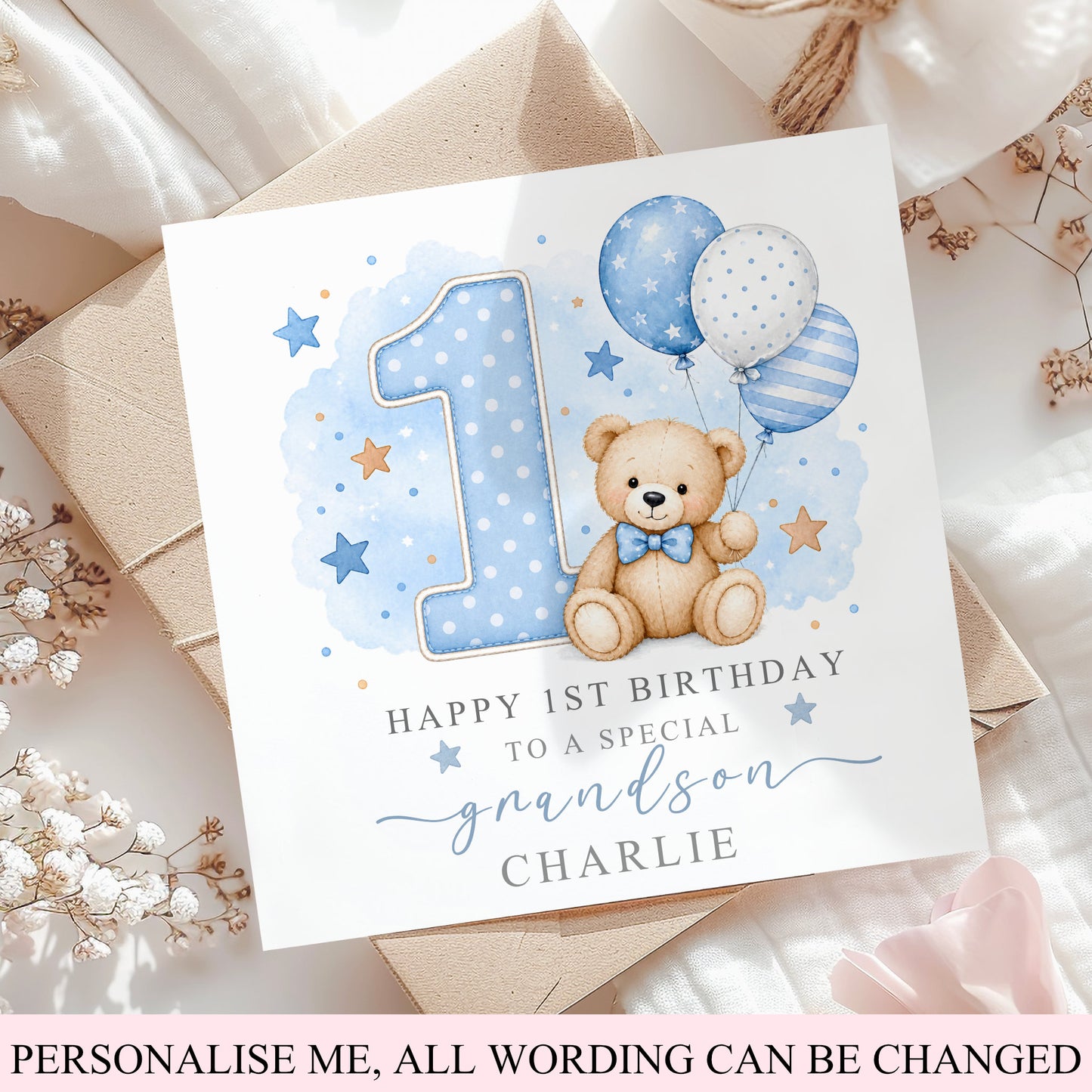 A customisable first birthday card for a grandson, featuring a watercolor teddy bear, balloons, stars, and a large number 1. The card is styled on top of a kraft paper envelope and white fabric with dried flowers.