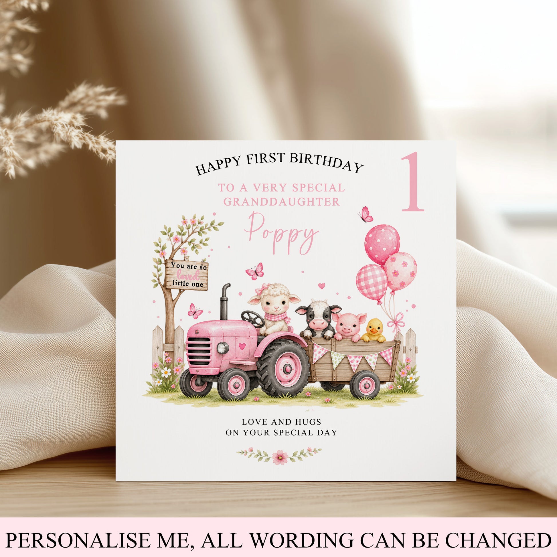 A customisable first birthday card for a granddaughter, featuring a pink tractor with cartoon animals, balloons, and a tree, displayed on a wooden surface with draped cream fabric.