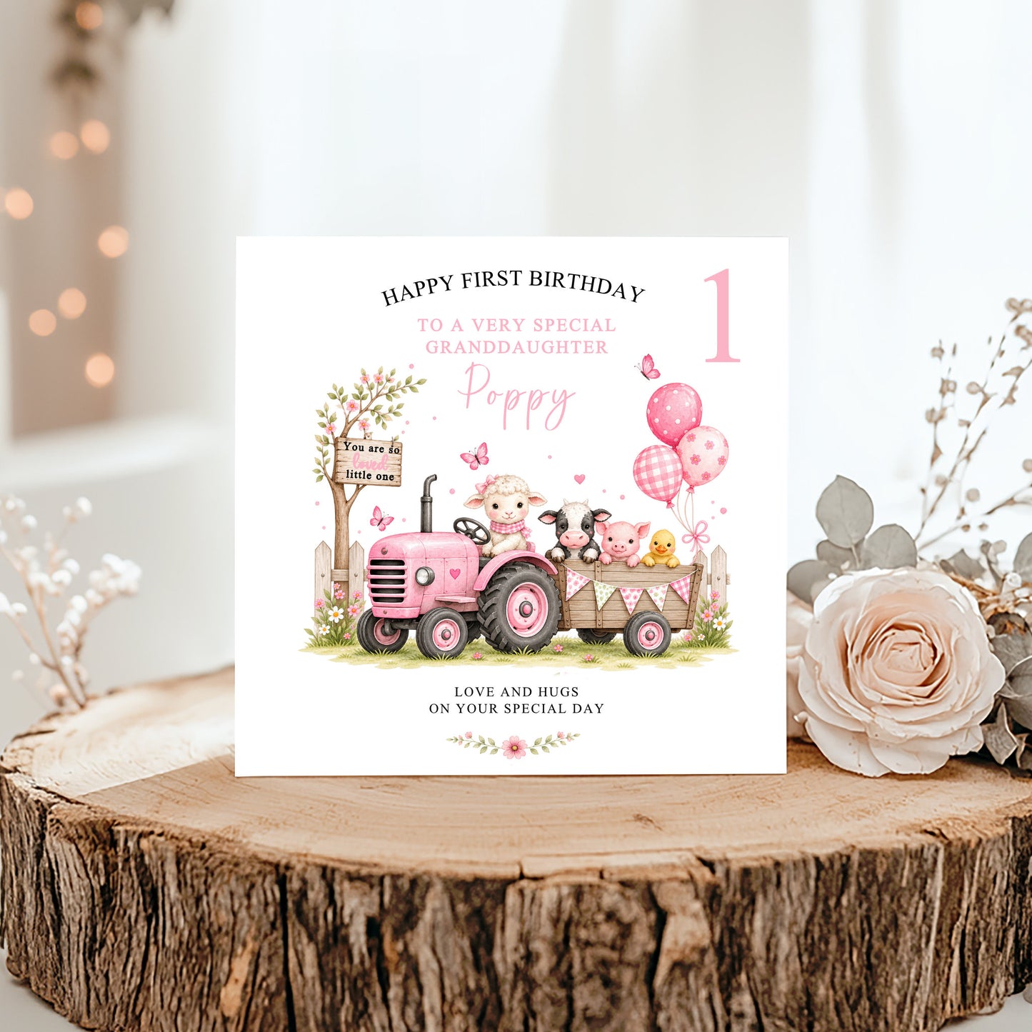A personalized first birthday card for a granddaughter, featuring a pink cartoon tractor and pig illustration, displayed on a rustic wood slice with a rose and dried flowers.