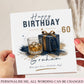 A personalized birthday card held by two hands. The card features a white background with a watercolor illustration of a whiskey glass and a blue gift box with a gold ribbon, alongside the text 'Happy BIRTHDAY 60' and the name 'Graham'.