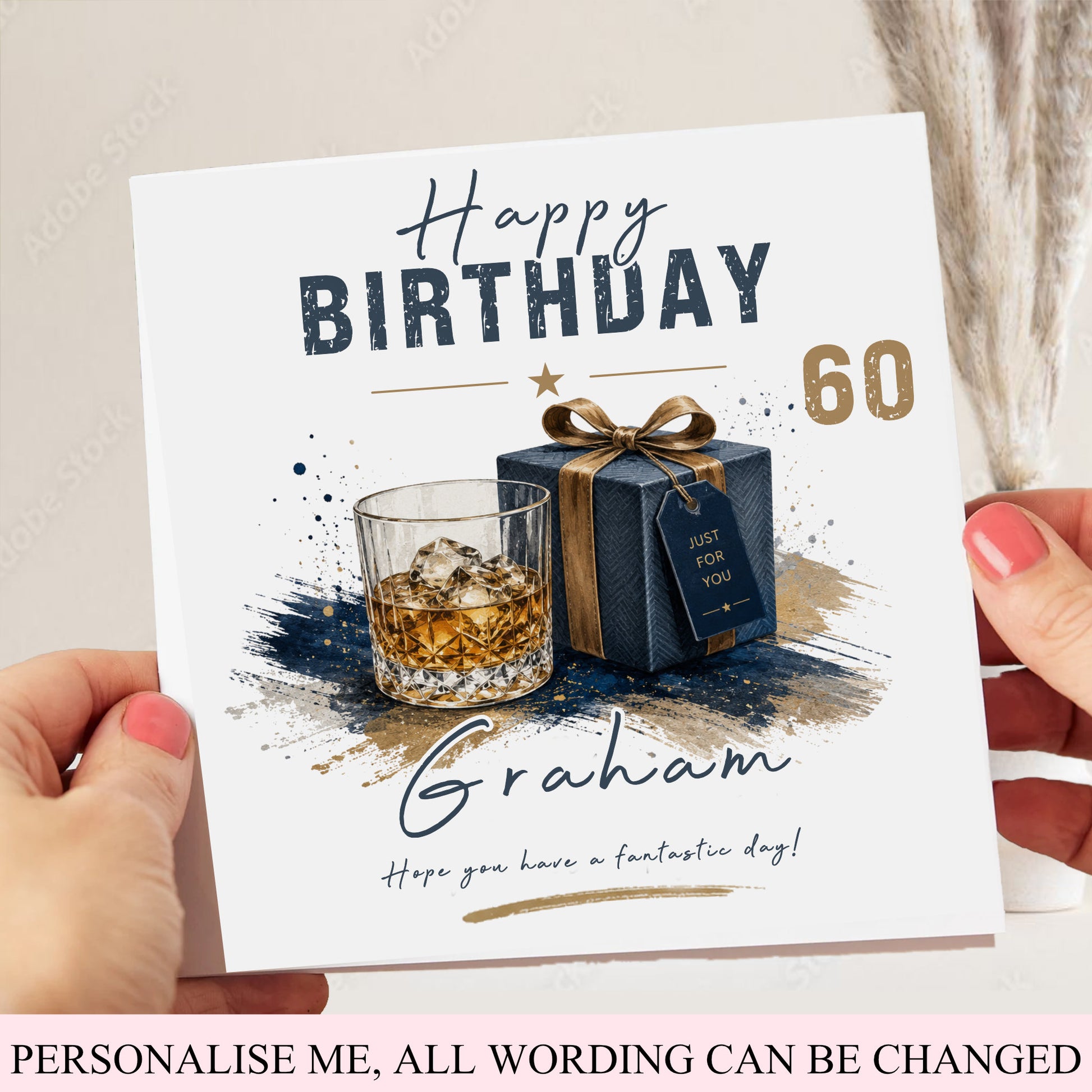 A personalized birthday card held by two hands. The card features a white background with a watercolor illustration of a whiskey glass and a blue gift box with a gold ribbon, alongside the text 'Happy BIRTHDAY 60' and the name 'Graham'.