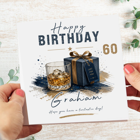 A personalized 60th birthday card held by two hands over a light wood background. The white card features a watercolor illustration of a whiskey glass, a navy blue gift box with a gold ribbon, and the text 'Happy Birthday 60' and the name 'Graham'.